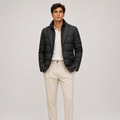 A man modeling a black quilted puffer jacket over a white polo shirt and cream trousers, standing against a light gray studio background.