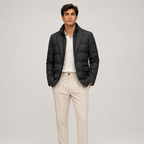 A man modeling a black quilted puffer jacket over a white polo shirt and cream trousers, standing against a light gray studio background.