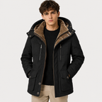 A man wearing a black hooded parka jacket with a brown sherpa-lined hood, paired with a black ribbed sweater and beige trousers, photographed against a light gray studio background.