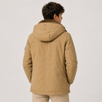 A man viewed from the back wearing a tan hooded jacket with a brown collar, paired with beige pants, against a plain light gray studio background.