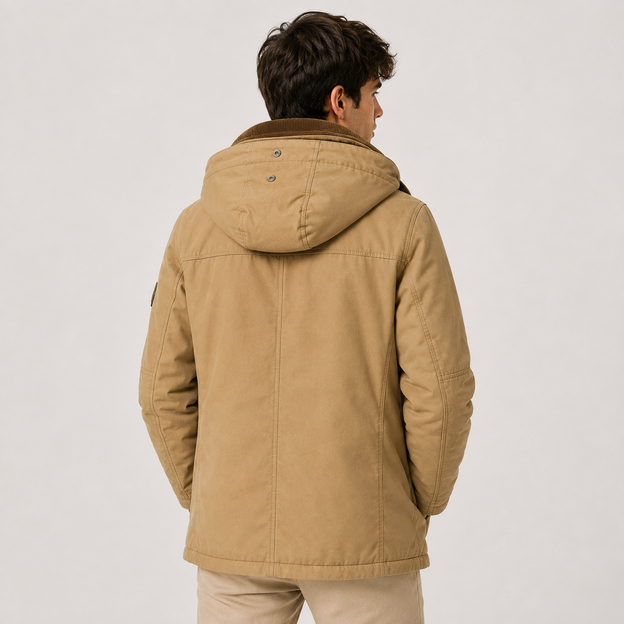 A man viewed from the back wearing a tan hooded jacket with a brown collar, paired with beige pants, against a plain light gray studio background.