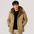 A man wearing a tan hooded winter parka jacket with a dark brown fleece-lined hood, layered over a black ribbed sweater and beige pants, photographed against a light gray studio background.