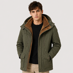 A man wearing an olive green hooded jacket with a brown sherpa-lined hood, layered over a black shirt and beige pants, photographed against a light gray studio background.