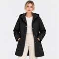 A woman modeling a black quilted hooded jacket with silver snap buttons, worn over a white top and cream trousers, against a plain light gray studio background.