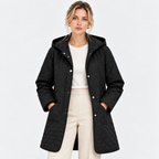 A woman modeling a black quilted hooded jacket with silver snap buttons, worn over a white top and cream trousers, against a plain light gray studio background.