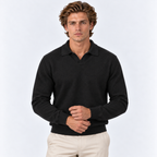 A man wearing a dark charcoal knit polo sweater and light beige trousers, standing against a light gray studio background.