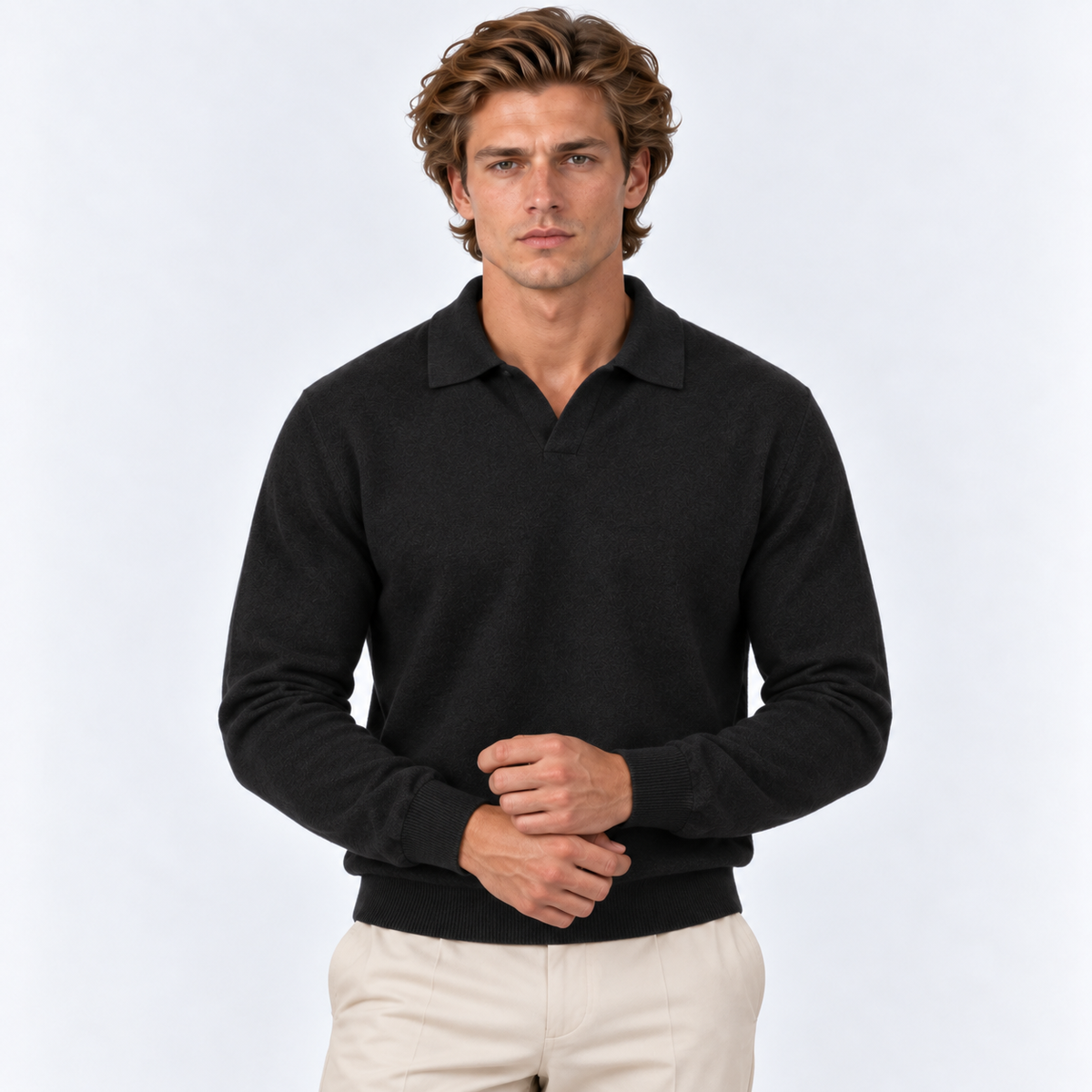 A man wearing a dark charcoal knit polo sweater and light beige trousers, standing against a light gray studio background.