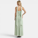 Back view of a woman wearing a sage green maxi dress with a lace-up back and flowing scarf detail against a light gray background.