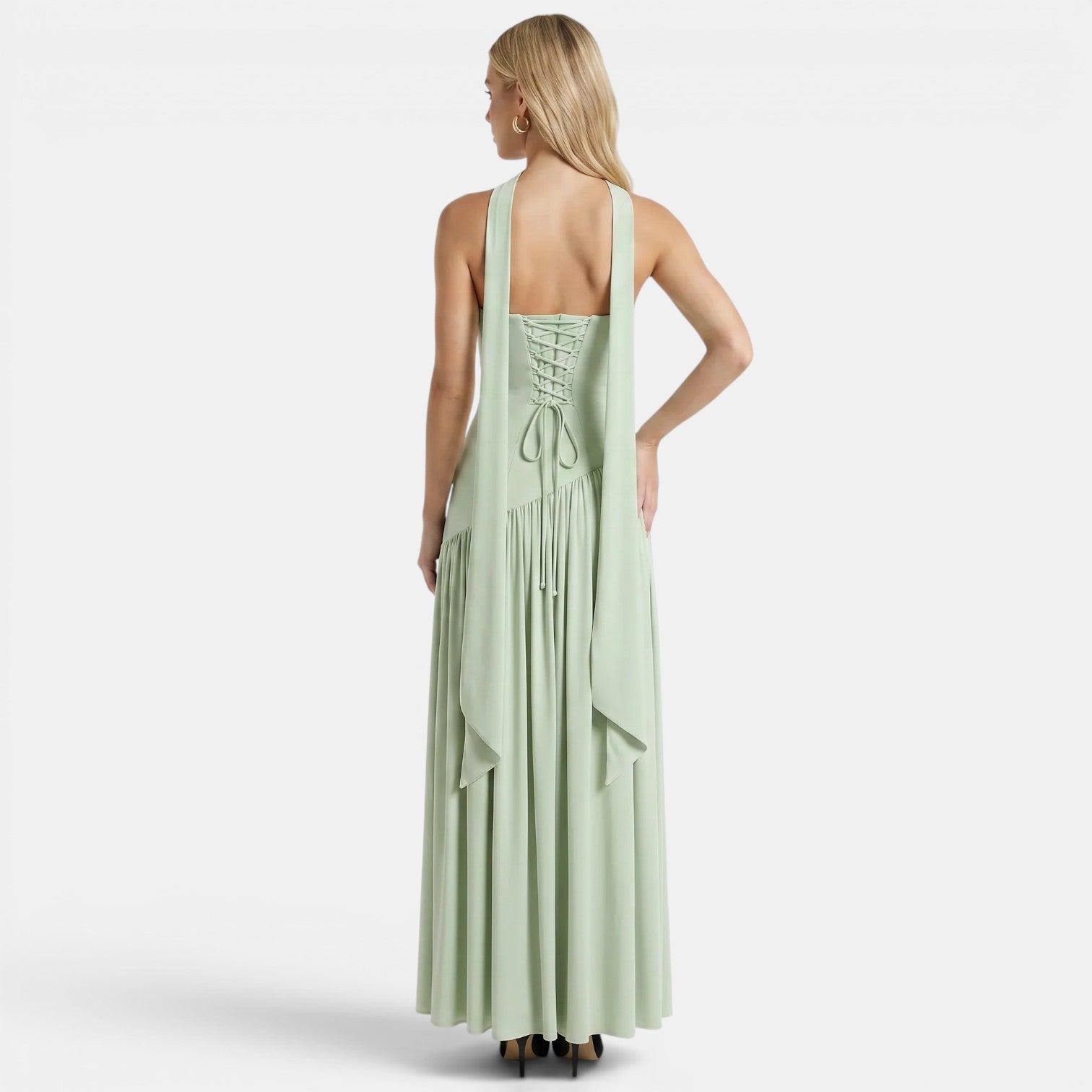 Back view of a woman wearing a sage green maxi dress with a lace-up back and flowing scarf detail against a light gray background.