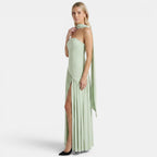 Side view of a woman wearing a sage green strapless maxi dress with a scarf neckline and thigh-high slit against a light gray background.