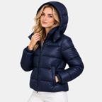 A woman wearing a navy blue hooded puffer jacket with white pants, photographed against a clean light gray studio background.