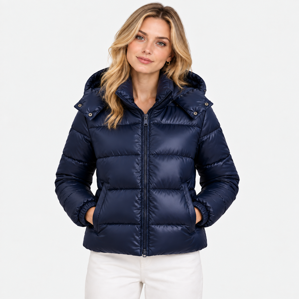 A woman wearing a navy blue hooded puffer jacket with a front zipper and horizontal quilting, paired with white pants, against a plain light gray background.