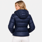 A woman shown from the back wearing a navy blue hooded puffer jacket and white pants against a light gray studio background.