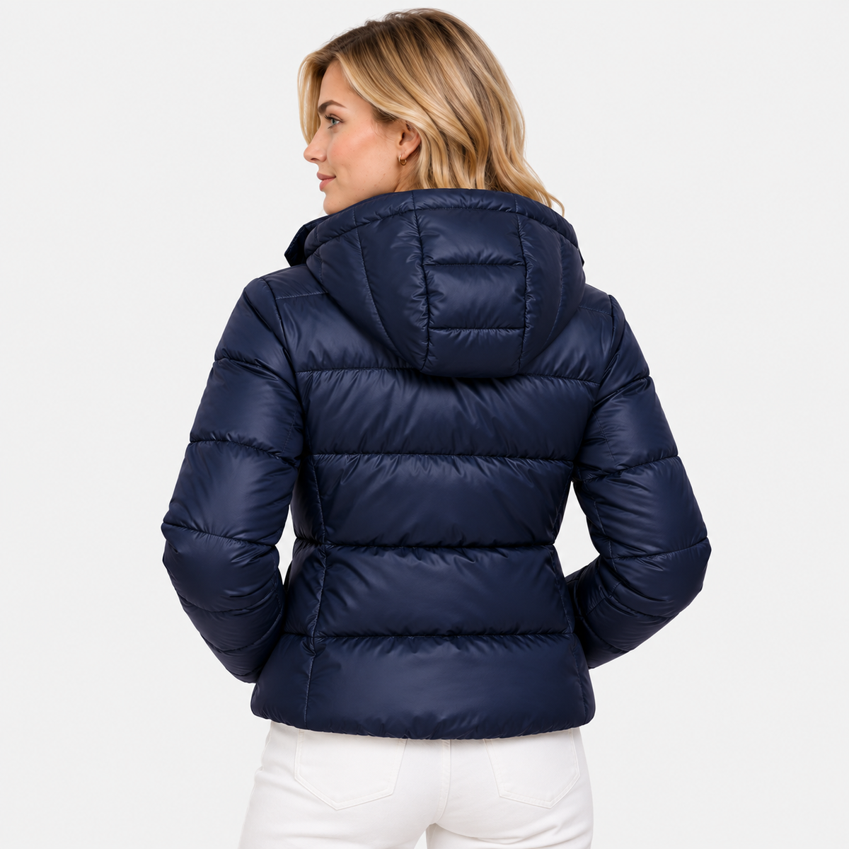 A woman shown from the back wearing a navy blue hooded puffer jacket and white pants against a light gray studio background.