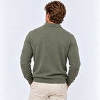 A man viewed from behind wearing an olive green knit polo sweater and light beige trousers, standing against a clean white studio background.