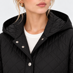 Close-up of a woman wearing a black quilted hooded jacket with silver snap buttons over a white ribbed top, against a light gray studio background.