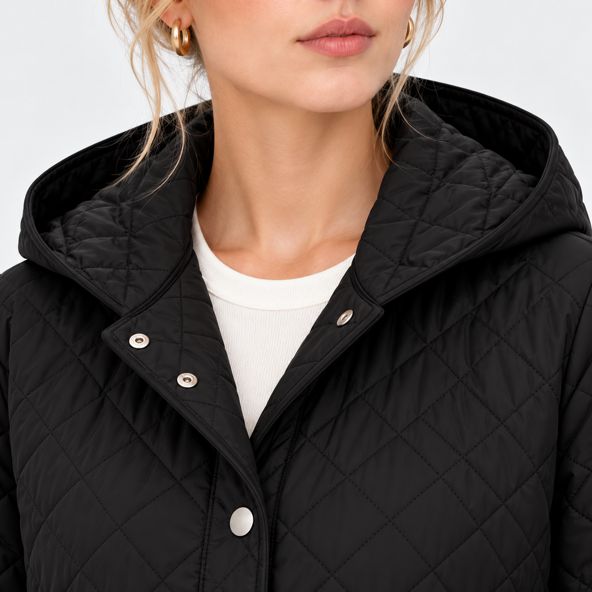 Close-up of a woman wearing a black quilted hooded jacket with silver snap buttons over a white ribbed top, against a light gray studio background.