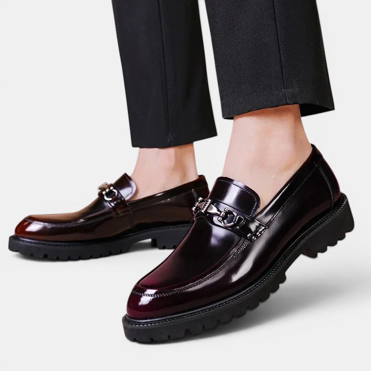Dark brown loafers with metal buckles worn with black pants on a light gray background