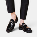 Black patent leather shoes with metal buckles worn with black pants on a light gray background