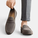Gray loafers being held up with a person wearing gray pants on a white background