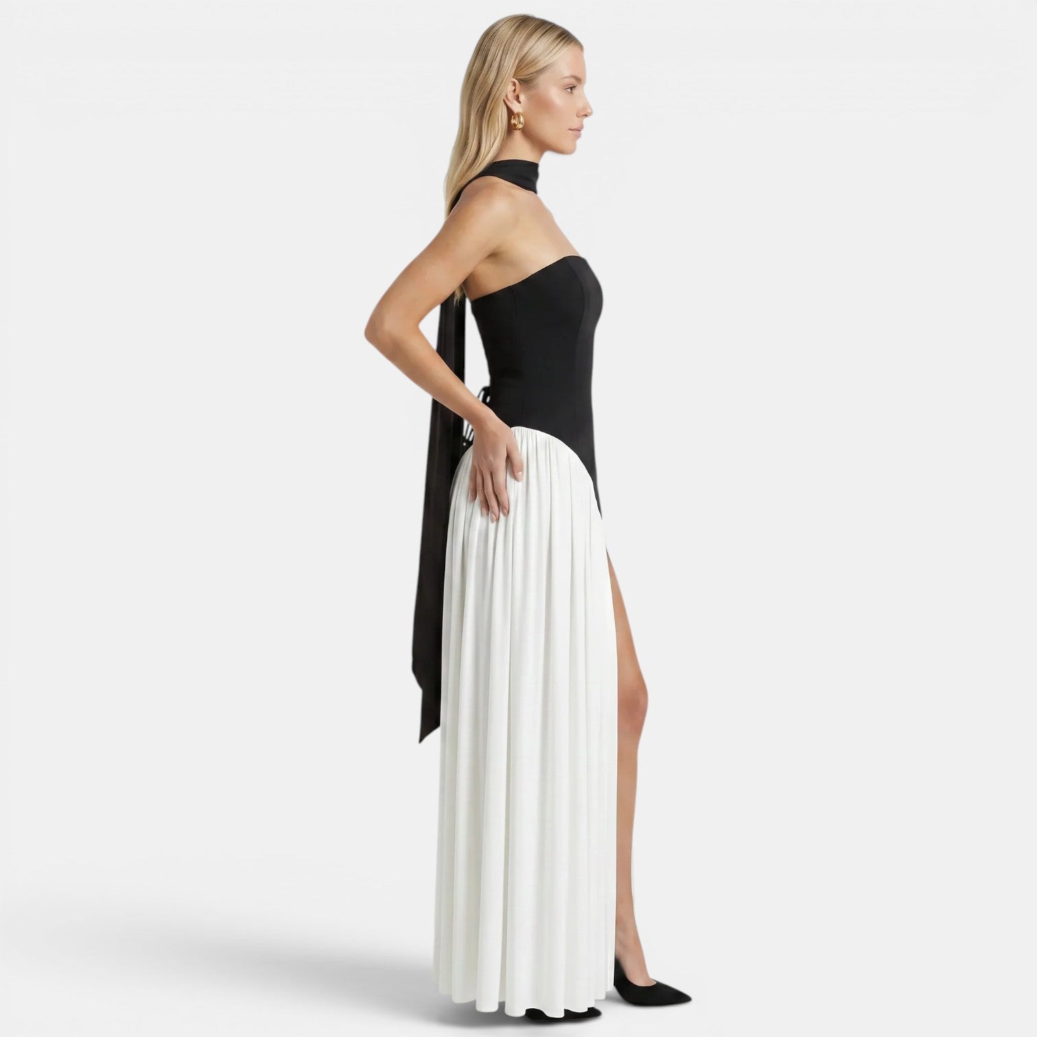 Side view of a woman wearing a black-and-white strapless maxi dress with a scarf neckline and thigh-high slit against a light gray background.