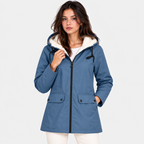 A woman wearing a blue hooded parka jacket with a cream sherpa-lined hood, black zipper, and front flap pockets, photographed against a light gray studio background.