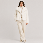 A woman modeling a white faux fur cropped jacket over a cream turtleneck and cream wide-leg trousers, standing against a light gray studio background.