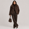 A model stands against a light gray studio background wearing a dark brown shaggy faux-fur jacket over a black turtleneck, paired with dark brown wide-leg trousers, holding a matching dark brown leather handbag.