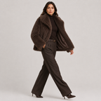 A woman models a dark brown faux fur jacket over a black turtleneck and dark brown wide-leg trousers, walking against a light gray studio background.