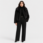 A model stands against a light gray studio background wearing a black faux fur jacket over a black turtleneck, paired with black wide-leg trousers and black pointed shoes.
