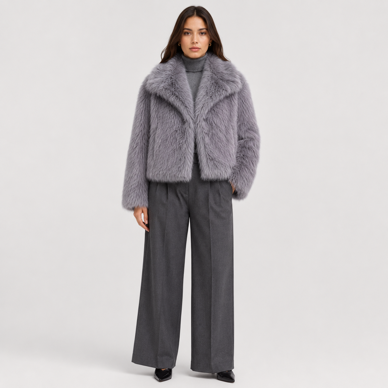 A model stands against a light gray studio background wearing a light purple-gray faux fur cropped jacket over a dark gray turtleneck and dark gray wide-leg trousers.