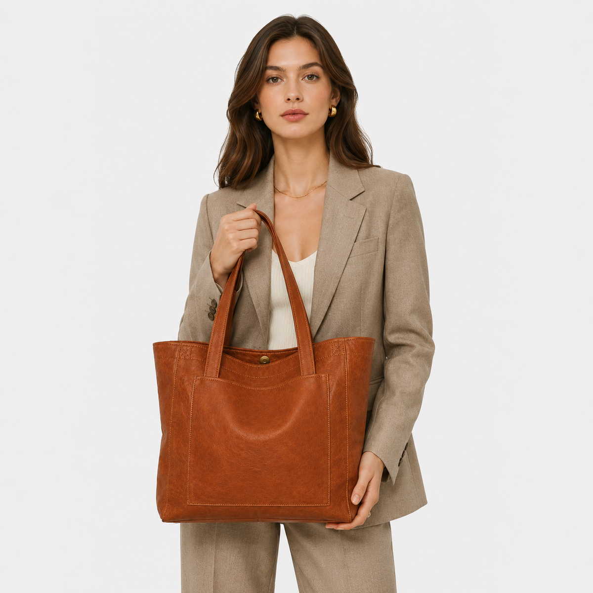 A woman wearing a beige blazer and matching trousers holds a large, distressed brown leather tote bag against a plain light gray background.