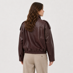 A woman shown from the back wearing a dark brown faux-leather bomber jacket with shoulder epaulets and a gathered elastic hem, paired with beige trousers, against a plain light gray studio background.