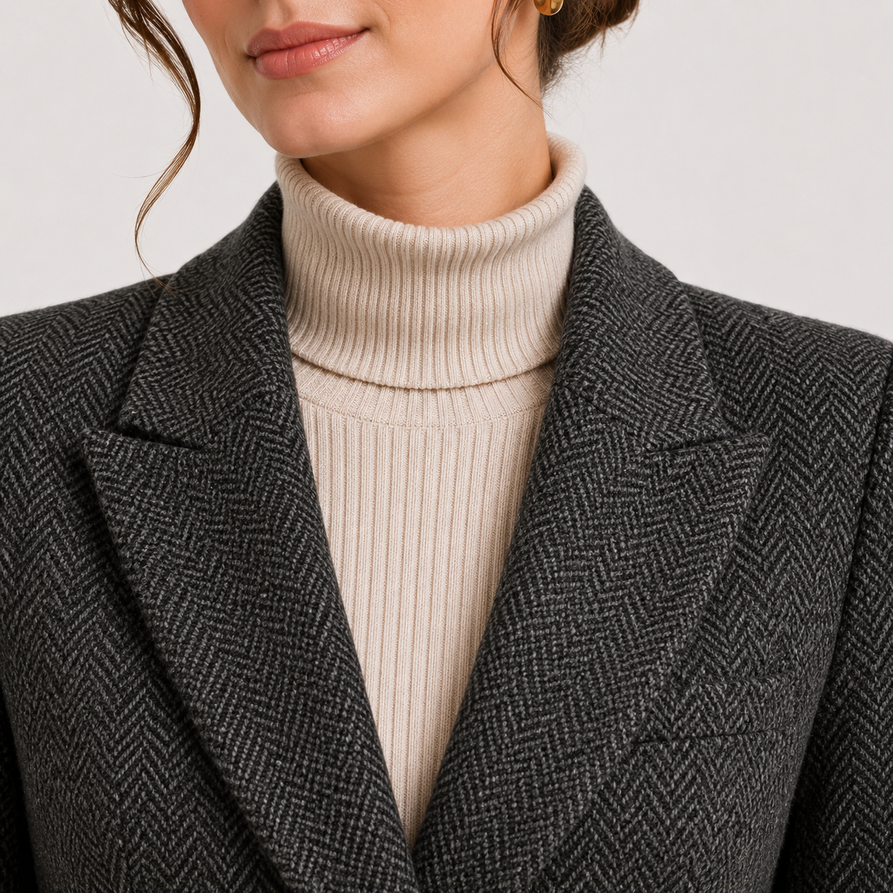 Close-up of a person wearing a dark gray and black herringbone blazer over a cream ribbed turtleneck sweater.