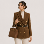 A woman models a brown double-breasted blazer with gold buttons, a black belt, and a matching brown handbag, styled over a cream turtleneck and cream trousers against a light gray studio background.