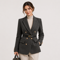 A woman models a dark gray double-breasted blazer with a matching waist belt, worn over a cream turtleneck and paired with beige trousers, holding a black handbag against a light gray studio background.