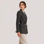 A woman modeling a dark gray textured double-breasted blazer with gold buttons, a black belt, and beige trousers, shown from a side profile against a light gray studio background.