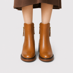 A pair of women's ankle boots in smooth tan-brown leather, shown on a model from the knees down. The boots feature side zippers with tassel pulls and a low block heel, set against a plain light gray background.