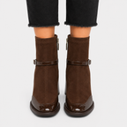 A pair of dark brown suede ankle boots with patent leather strap details and side zippers, shown on a model wearing black frayed-hem jeans against a light gray background.