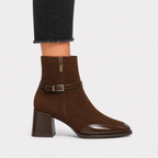 A brown suede ankle boot with a block heel, glossy toe cap, and a side buckle strap, shown on a model's foot against a light gray background.