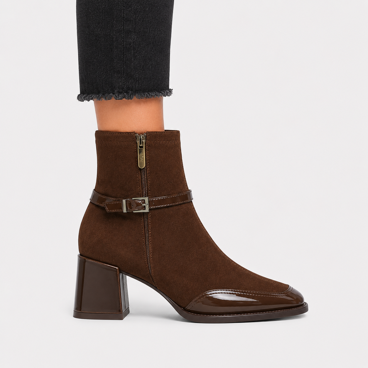 A brown suede ankle boot with a block heel, glossy toe cap, and a side buckle strap, shown on a model's foot against a light gray background.