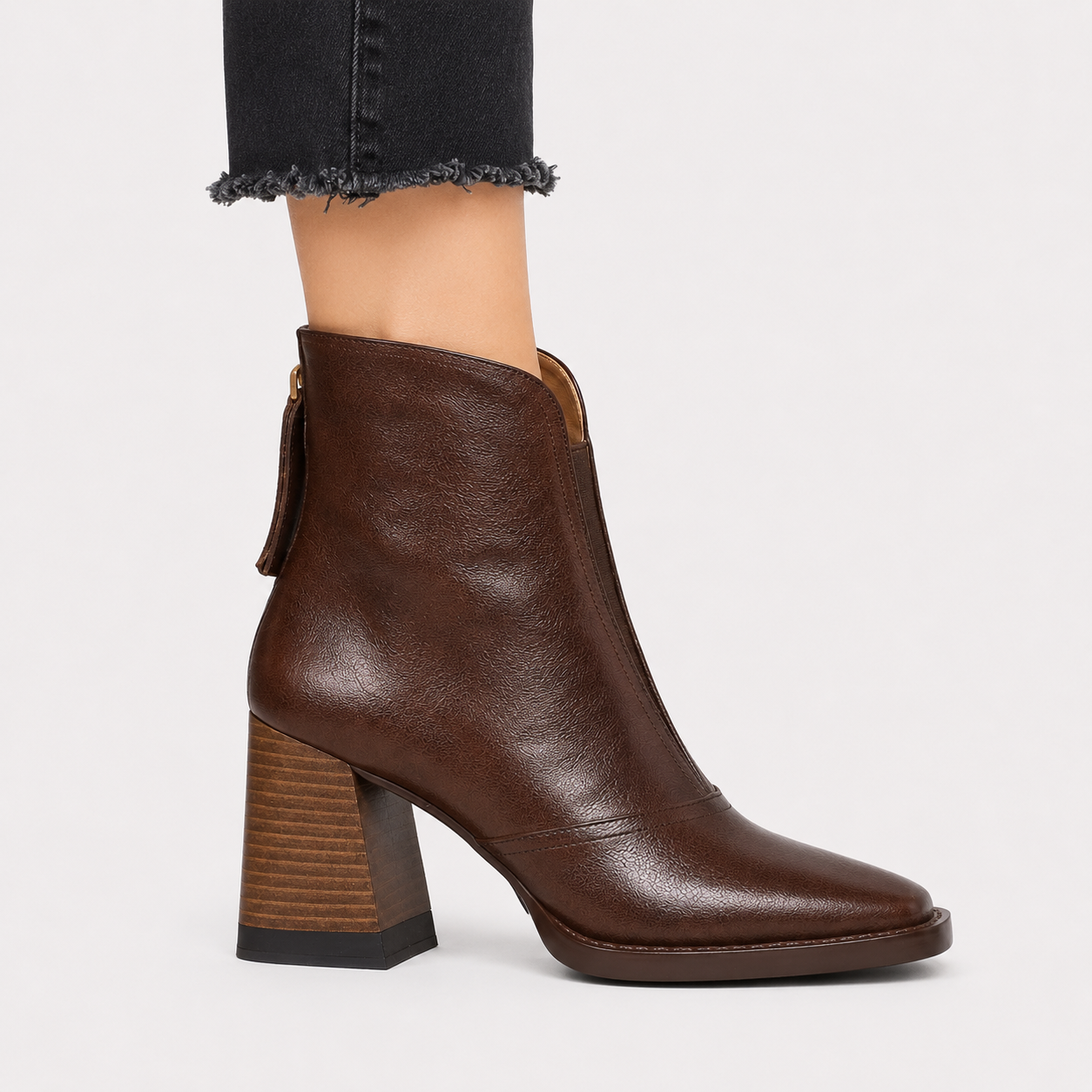 A dark brown leather ankle boot with a chunky block heel, shown on a model's foot with frayed black jeans against a light gray background.
