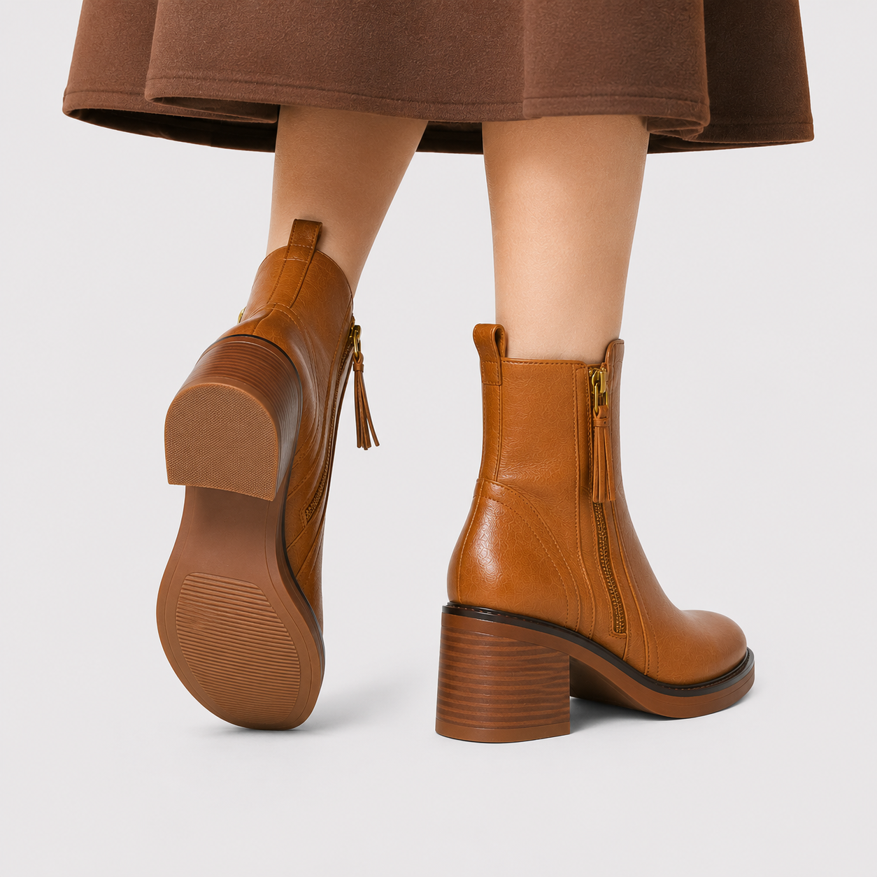 A pair of women's ankle boots in tan brown leather, shown on a model from the back and side. The boots feature a chunky block heel, a side zipper with a tassel pull, and a low block heel.