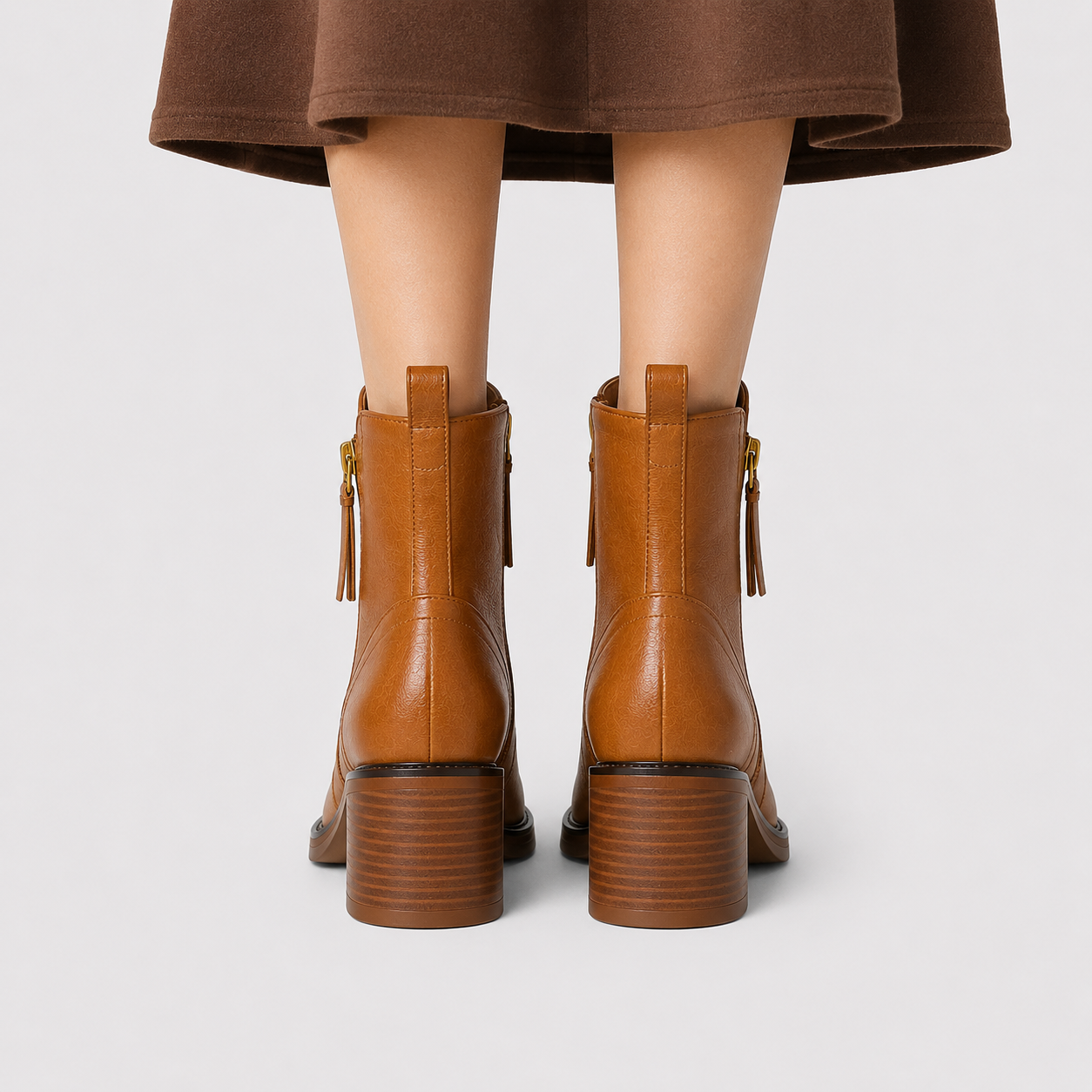 Back view of a person wearing brown ankle boots with block heels and gold-tone zipper pulls, shown against a light gray studio background.