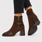 A pair of women's brown suede ankle boots with glossy patent leather block heels, side zippers, and decorative buckle straps, shown on a model wearing black jeans against a light gray background.