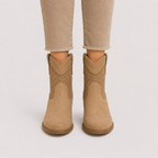 A pair of tan suede western-style ankle boots with perforated side panels, shown on a model wearing beige cropped jeans against a light gray studio background.