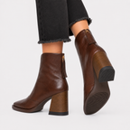 A pair of dark brown leather ankle boots with block heels, shown on a model wearing black frayed-hem jeans against a light gray background.