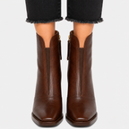 A pair of dark brown leather ankle boots with side zippers, shown on a model wearing black frayed-hem jeans against a light gray background.