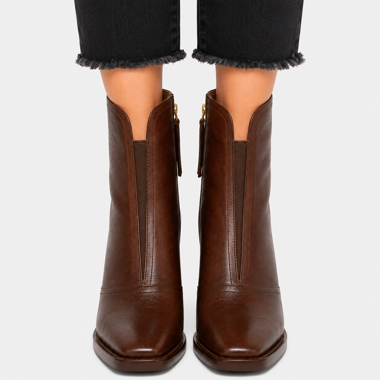 A pair of dark brown leather ankle boots with side zippers, shown on a model wearing black frayed-hem jeans against a light gray background.