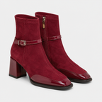 A pair of women's ankle boots in deep burgundy suede with a glossy patent leather toe cap, block heel, and a decorative side buckle strap, shown against a light gray background.